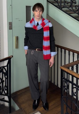 Joven de pie en interiores junto a una barandilla de escaleras, vistiendo pantalones grises, suéter negro con hombros descubiertos sobre camisa azul y una bufanda de rayas rojas y azules.