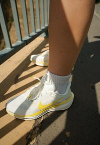 Baltos Nike bėgimo bateliai su geltonais akcentais ir tinklelio tekstūra. Dėvimi su baltais kojiniais, matomi ant akmenimis grįsto paviršiaus po turėklu.
