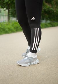 Zwarte sportleggings met witte drie-strepen details aan de zijkanten, gecombineerd met grijze hardloopschoenen en witte enkelkousen.