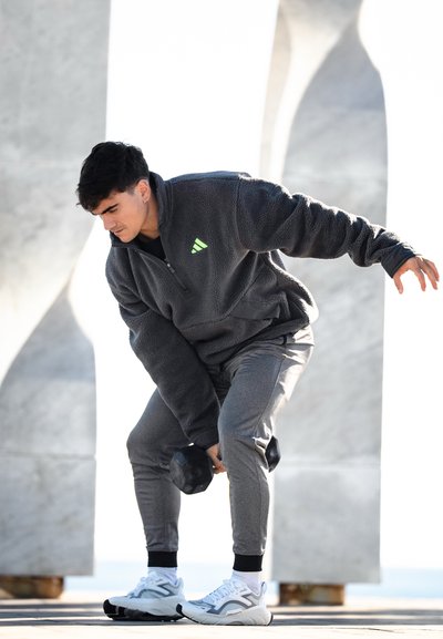 Sudadera gris de forro polar con media cremallera y logo verde, combinada con pantalones jogger grises ajustados y zapatillas blancas. Sosteniendo una pesa durante el ejercicio.