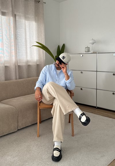Camisa abotonada azul claro, pantalones beiges, zapatos negros y una gorra. Silla de madera y sofá beige en una habitación luminosa con plantas y cortinas.