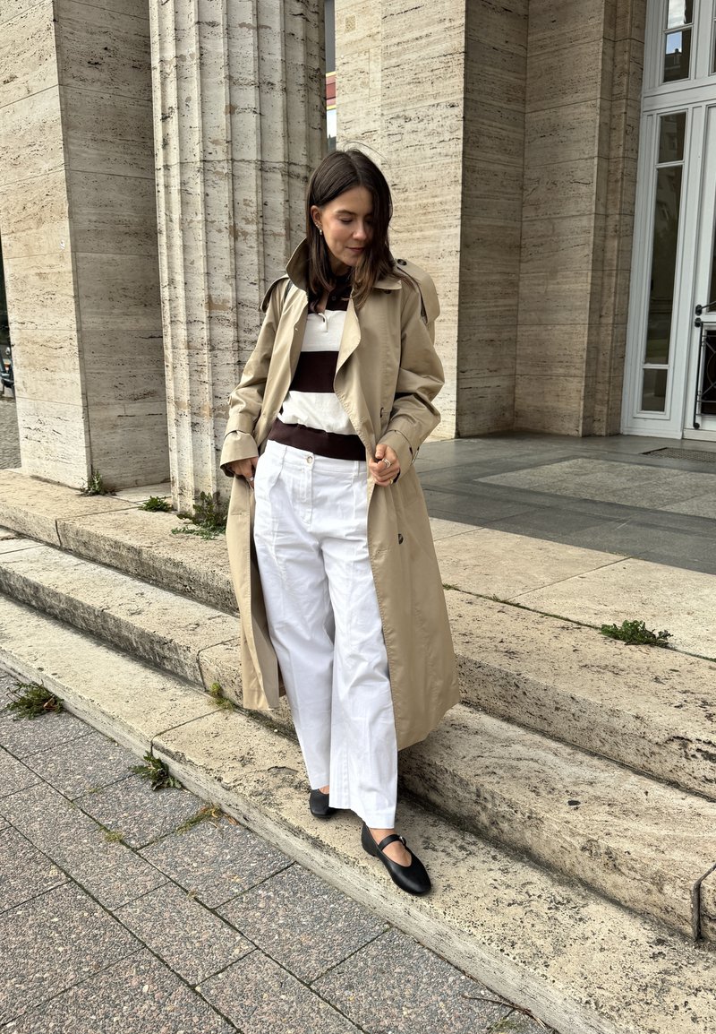 Cappotto beige con ampi revers, top a righe bianco e marrone, pantaloni bianchi a gamba larga e scarpe basse nere su gradini di pietra.