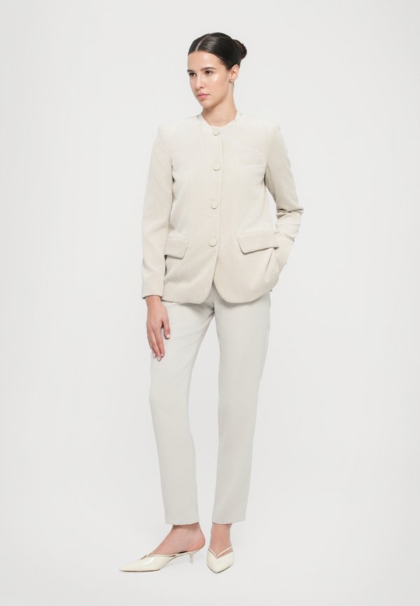 Blazer de corduroy color beige claro con cuello redondo, dos bolsillos frontales, combinado con pantalones ajustados gris claro y mules blancos de punta.