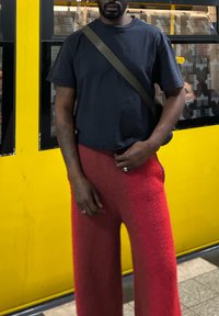 T-shirt noir à manches courtes associé à un pantalon rouge texturé et ample. La personne tient un sac et se tient devant un fond jaune vif.