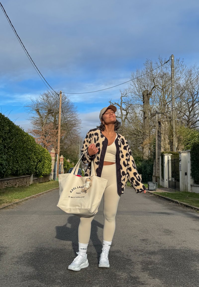 Femme portant une tenue de sport beige, un cardigan à imprimé léopard et une casquette, marchant dans la rue en portant un grand sac cabas en toile en plein jour.