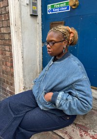 Denim bomberjakke med ribkanter, båret over mørkeblå jeans. Lysebrunt hår bundet med en scrunchie, runde briller og geometriske øreringe.