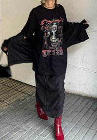 Černé oversized tričko s grafickým designem a červenými akcenty, kombinované s vícevrstvovou černou sukní a lesklými červenými kotníkovými botami.