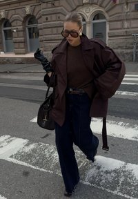 Overdimensjonert brun trenchcoat over en brun genser, kombinert med mørkeblå vidbukser og svarte hansker, holding en svart håndveske.