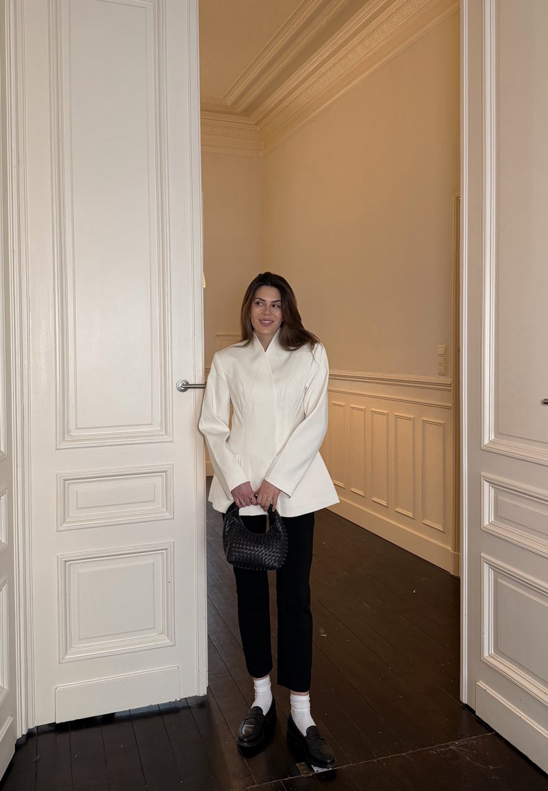 Donna con giacca bianca su misura, pantaloni neri, calzini bianchi, mocassini neri, che tiene una borsa intrecciata nera, in piedi nella porta di una stanza color crema.