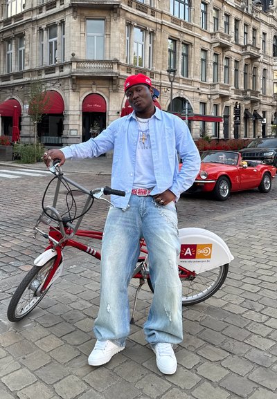 Bicicleta roja con acentos blancos, persona con camisa azul claro y jeans desgastados, gorra roja, entorno urbano de adoquines con edificios.