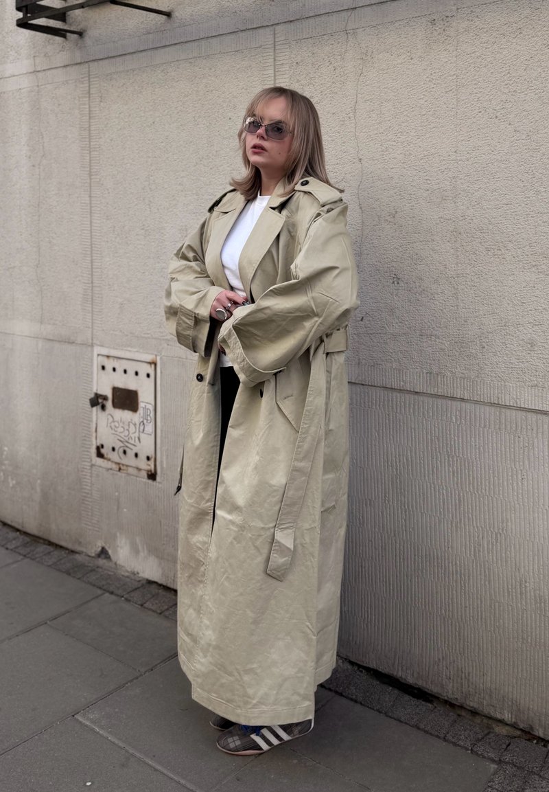 Femme portant un trench-coat beige oversize, une chemise blanche, des lunettes de soleil et des baskets, debout contre un mur urbain texturé.