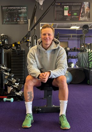 Sudadera gris, pantalones cortos negros y zapatillas deportivas verdes. Sentado en un banco de pesas con mancuernas y equipo de fitness en un gimnasio.