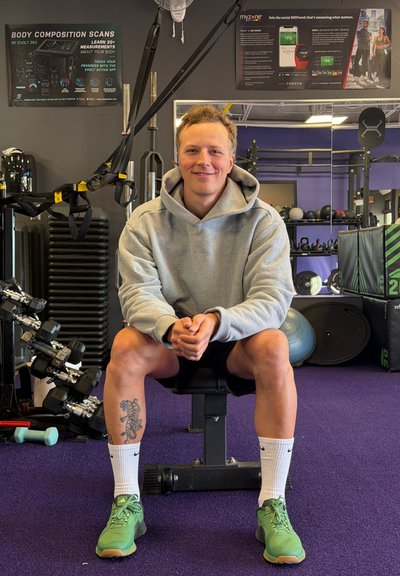 Sudadera gris, pantalones cortos negros y zapatillas deportivas verdes. Sentado en un banco de pesas con mancuernas y equipo de fitness en un gimnasio.