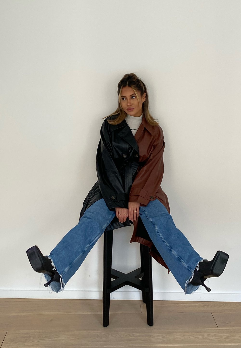 Zwart en bruin twee-tonen oversized trenchcoat, blauwe distressed wijde jeans en zwarte plateauhakken, zittend op een zwarte kruk.