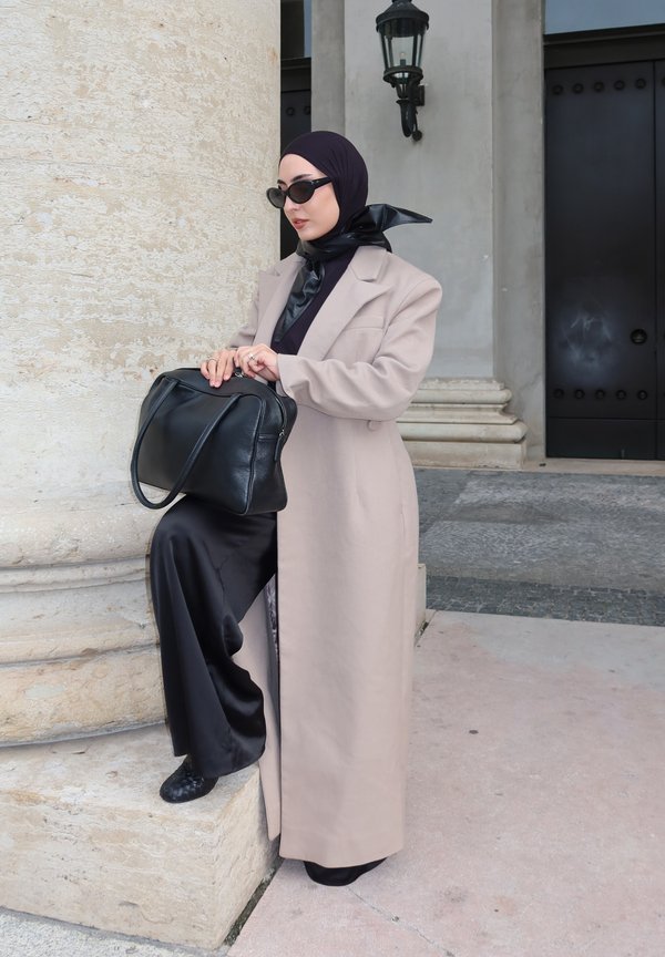Une femme portant un hijab foncé et des lunettes de soleil, vêtue d’un manteau beige, tient un sac à main noir près d’une colonne en pierre, à l’extérieur d’un bâtiment.
