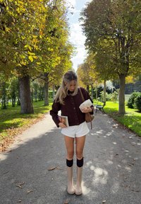 Ruda mygtukų megzta kardiganas, baltos šortai, šviesiai rudos iki kelių batai su tamsiais apvadais; laiko knygas ir mažą kuprinę lauko aplinkoje.