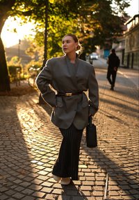 Sivý oversize blazer s vysokým golierom, zatužený v páse. Kombinovaný s čiernymi pruhovanými širokými nohavicami a bielymi platformovými topánkami. Čierna kabelka.