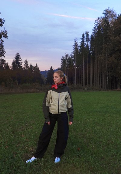 Chaqueta ligera bicolor en beige y verde oscuro, con capucha y cremalleras, combinada con pantalones negros holgados y zapatillas blancas.