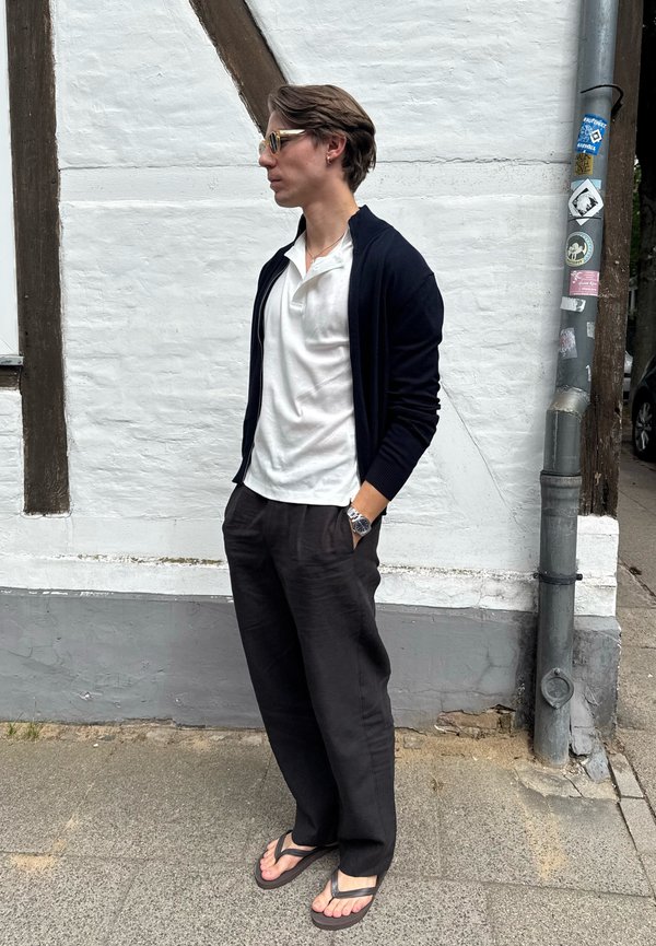 Jonge man met zonnebril, wit shirt, zwarte cardigan, zwarte broek en slippers, staand op het trottoir naast een witte muur en een paal.