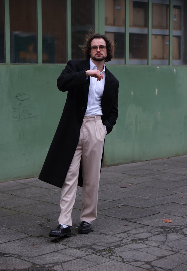 Schwarzer Wollmantel, weißes Hemd, beige Hose und glänzend schwarze Schuhe. Steht auf dem Gehweg vor einer grünen Wand.