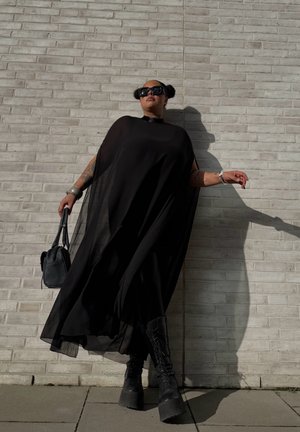 Vestido de capa negro transparente con un cuello alto y mangas largas, combinado con botas negras de plataforma gruesa y un bolso negro pequeño.