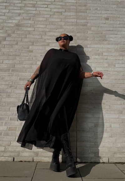 Vestido de capa negro transparente con un cuello alto y mangas largas, combinado con botas negras de plataforma gruesa y un bolso negro pequeño.