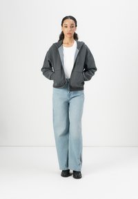 Grå zip-hoodie med dragsko i huvan, tillsammans med en vit t-shirt och ljusblå vida jeans med en subtil svart design nära fållen.