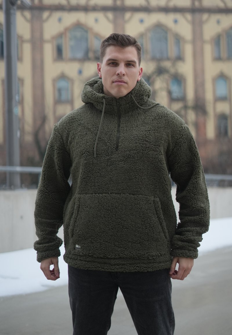 Jeune homme debout en plein air portant un sweat à capuche en polaire verte, avec un sol enneigé et un bâtiment flou en arrière-plan.