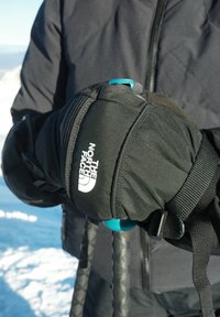 Gants isolants noirs avec un logo blanc, tenant des bâtons de trekking avec des poignées texturées noires et des accents bleus sur les manches, sur fond de neige.