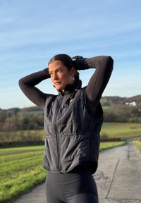 Fekete puffertestű mellény magas gallérral, steppelt mintával és elülső cipzárral. Hosszú ujjú fekete felső fölött viselve, zöld táj hátterében.