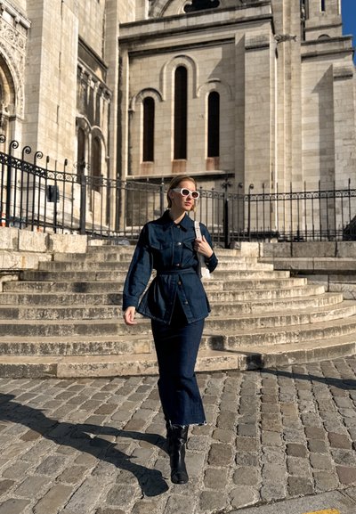 Chaqueta de mezclilla con botones frontales, cintura ceñida con cinturón, y dos bolsillos, combinada con una falda de mezclilla. Las botas negras hasta la rodilla y las gafas de sol blancas completan el look.