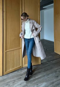 Krémszínű trench coat, dupla soros gombokkal, fehér bordázott garbóval és kék, testhezálló farmernadrággal kombinálva, feket szegélyű bokacsizmával kiegészítve.