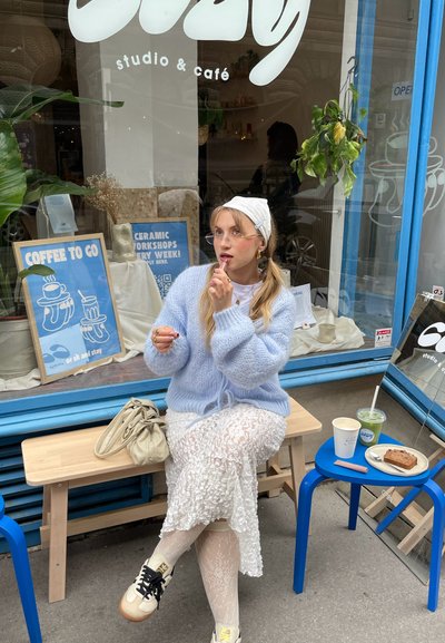 Suéter de lana azul claro, falda de encaje blanca, planta colgante, banco de madera, mesa azul con café, pastelería y bebida verde. Ventana de cafetería al fondo.