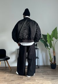Jachetă bomber pufosă neagră cu manșete ribbate, purtată peste o cămașă albă. Pantaloni negri largi și o căciulă tricotată neagră. Decor interior cu plante.