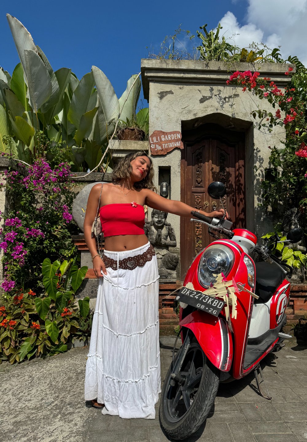 Rode scooter met een zilveren voorplaat, geparkeerd op een stenen pad. Op de achtergrond zijn groene bladeren en roze bloemen te zien, met een decoratieve muur.