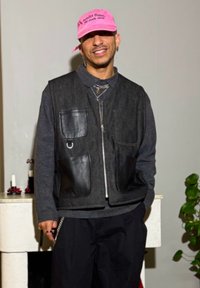 Veste noire et grise sans manches avec plusieurs poches, portée par-dessus une chemise bleu marine, associée à un pantalon foncé et une casquette rose.