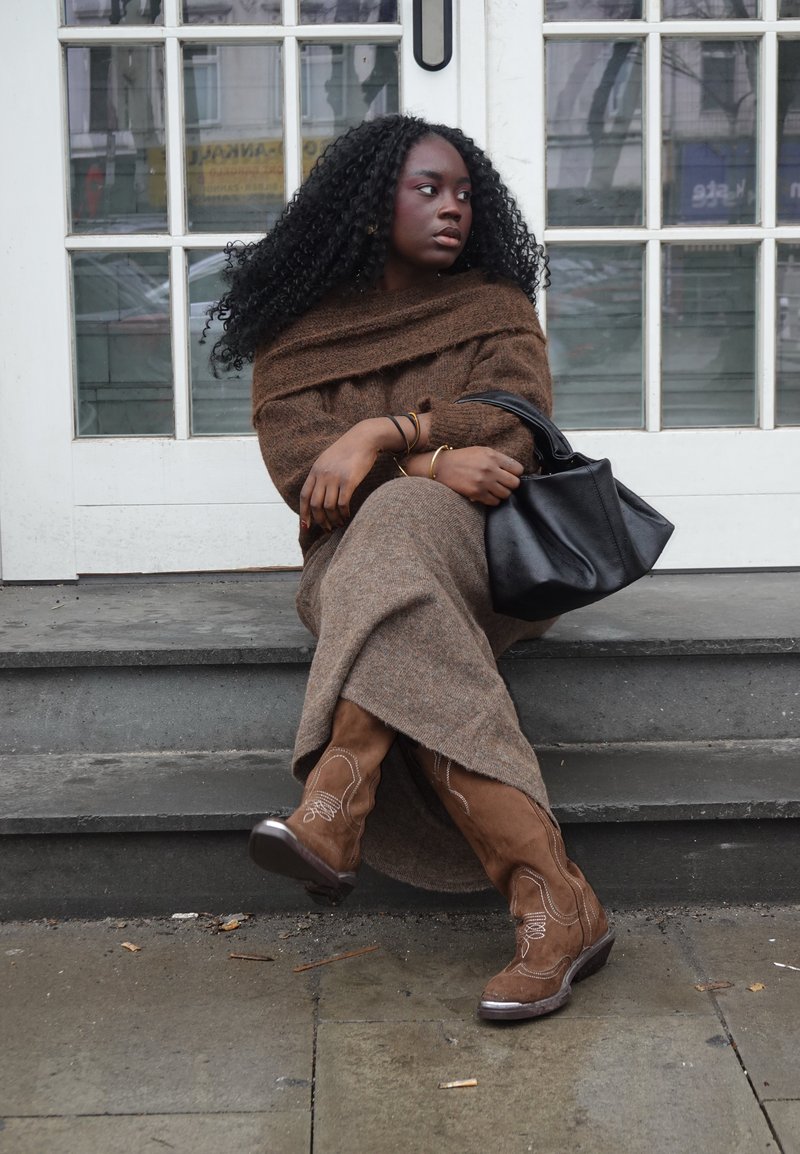 Femme aux cheveux bouclés, vêtue d'une robe marron et de bottes, assise sur des marches, tenant un sac à main noir, regardant sur le côté devant une porte à vitres.