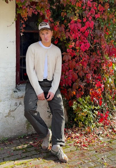 Cardigan beige sobre una camisa blanca, pantalones cargo oscuros y zapatillas beige. El fondo presenta enredaderas rojas y verdes contra una pared blanca.