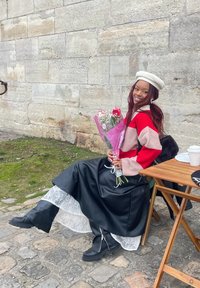 Rdeče in roza prugasta pulover, črna umetna usnjena krilo z čipkastim robom, črne kolenske škornje, drži roza vrtnice, sedi poleg lesenega mize.