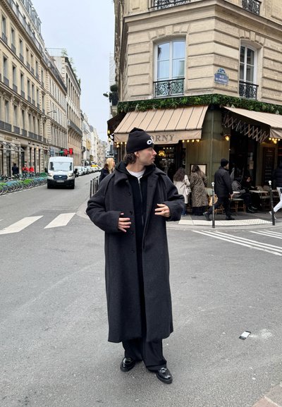 Abrigo largo de lana negra con un corte relajado, cierres de botones y un cuello ancho. Llevado sobre una camisa negra y pantalones oscuros con zapatos brillantes.