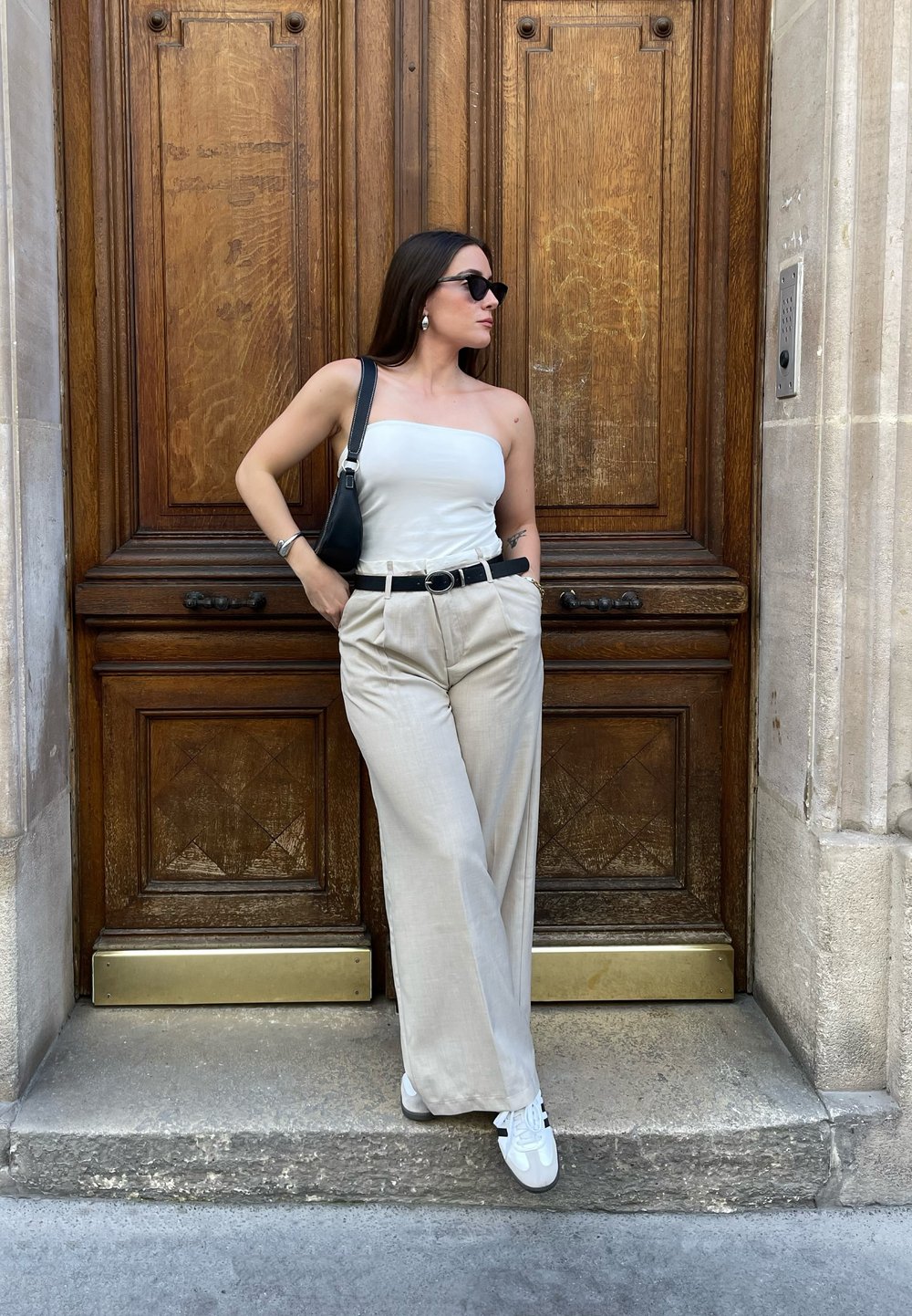 Λευκή τοπ χωρίς straps, μπεζ φαρδιές παντελόνες με μαύρη ζώνη, μαύρη τσάντα και λευκά αθλητικά παπούτσια. Στηρίχθηκε μπροστά σε μια ξύλινη πόρτα.