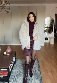 Femme portant un manteau en fausse fourrure blanche, un col roulé bordeaux, une mini-jupe en cuir, des bottes hautes jusqu'aux genoux, tenant un sac à main bordeaux dans un salon.