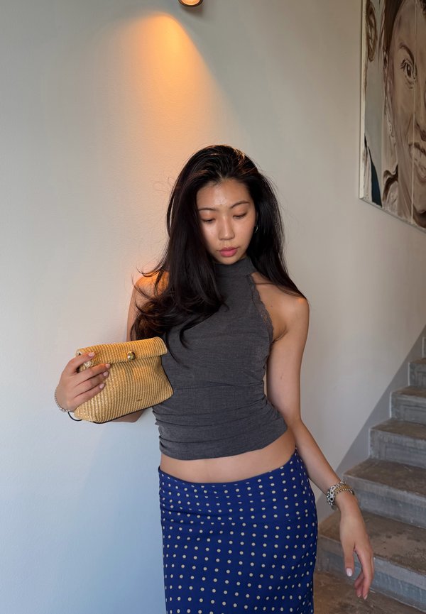 Donna con lunghi capelli scuri che indossa una canotta grigia e una gonna blu a pois, che tiene una piccola pochette intrecciata vicino a una scala.