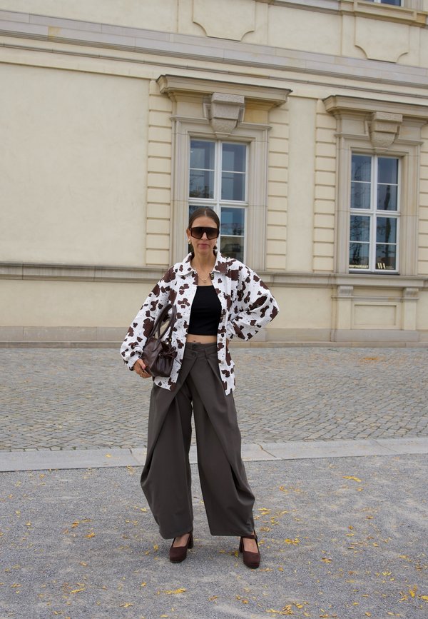 Mujer que lleva una chaqueta extragrande con estampado de vaca, top corto negro, pantalones anchos marrones, tacones marrones y gafas de sol, de pie en una calle empedrada cerca de un edificio beige.
