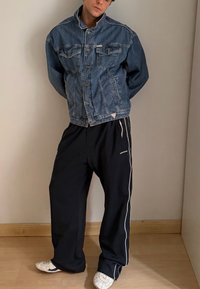 Giacca di denim con design a doppia tasca sul petto, colore blu medio. Pantaloni sportivi neri larghi con strisce bianche ai lati; scarpe sportive bianche.