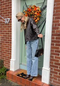 Brown leather jacket, light blue straight-leg jeans, black heeled boots, and a black handbag on a front porch adorned with a floral wreath.