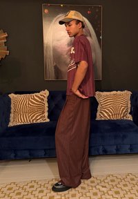 Νεαρός άνδρας με μπεζ καπέλο, πουκ άμισο με μπορντό λεπτές ρίγες και φαρδιές καφέ παντελόνες στέκεται πλάγια σε ένα σαλόνι με σκούρο μπλε καναπέ και μαξιλάρια με σχέδια.