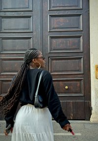 Maglione lungo nero a maniche, gonna bianca a strati, capelli intrecciati, orecchini rotondi, tiene il rossetto, in piedi davanti a una grande porta di legno.