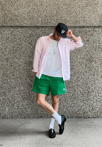 Conjunto masculino que incluye una camisa de botones a rayas de color rosa claro, una camiseta blanca, pantalones cortos verdes, calcetines blancos y mocasines negros. Gorra negra con logo.