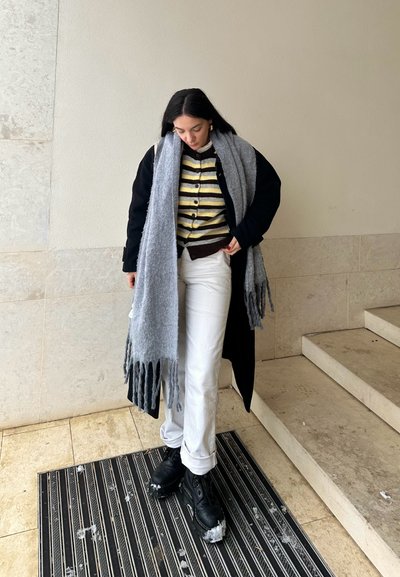 Bufanda con flecos gris sobre un abrigo negro, suéter a rayas marrón y amarillo, pantalones blancos y botas negras con nieve. De pie sobre una alfombra a rayas.